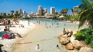 Een strand met mensen die zwemmen en zonnebaden, een stadsachtergrond in de verte, en grote rotsen in de buurt van het water.