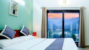 Een hotelkamer met een bed, nachtkastje, rode lamp en groot raam met uitzicht op bergen en een zonsondergang.