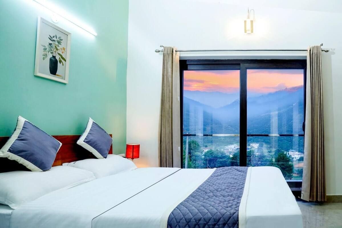 A hotel room with a bed, a nightstand, a red lamp, and a large window with a view of mountains and a sunset.