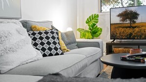 A modern living room with a grey sofa, patterned cushions, a large green plant, a television displaying a field, and a coffee table with a book. - Spacious 3-Bed House in Leafy Paddington (Paddington)