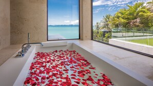 Une baignoire remplie de pétales de rose, une grande fenêtre donnant sur une plage et des montagnes, et un design moderne pour la salle de bain.