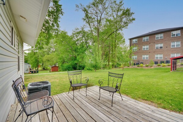 A wooden deck with two metal chairs and a grill, overlooking a grassy yard with trees and a brick building in the background.