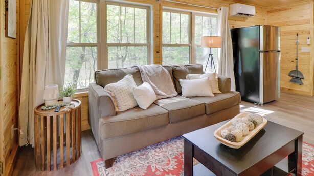 Ein gemütliches Blockhauss-Interieur mit einer Couch, einem Esstisch, einem Kühlschrank und einem großen Fenster mit Blick auf Bäume.