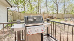 Una parrilla exterior con pantalla digital y tres perillas de control, ubicada en una terraza con valla de madera y árboles al fondo.