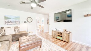 Un salón amplio con ventilador de techo, un televisor de pantalla plana grande, un mueble de madera y un sofá con cojines estampados.