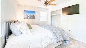 Un dormitorio con cama, ventilador de techo, televisor montado y ventana.