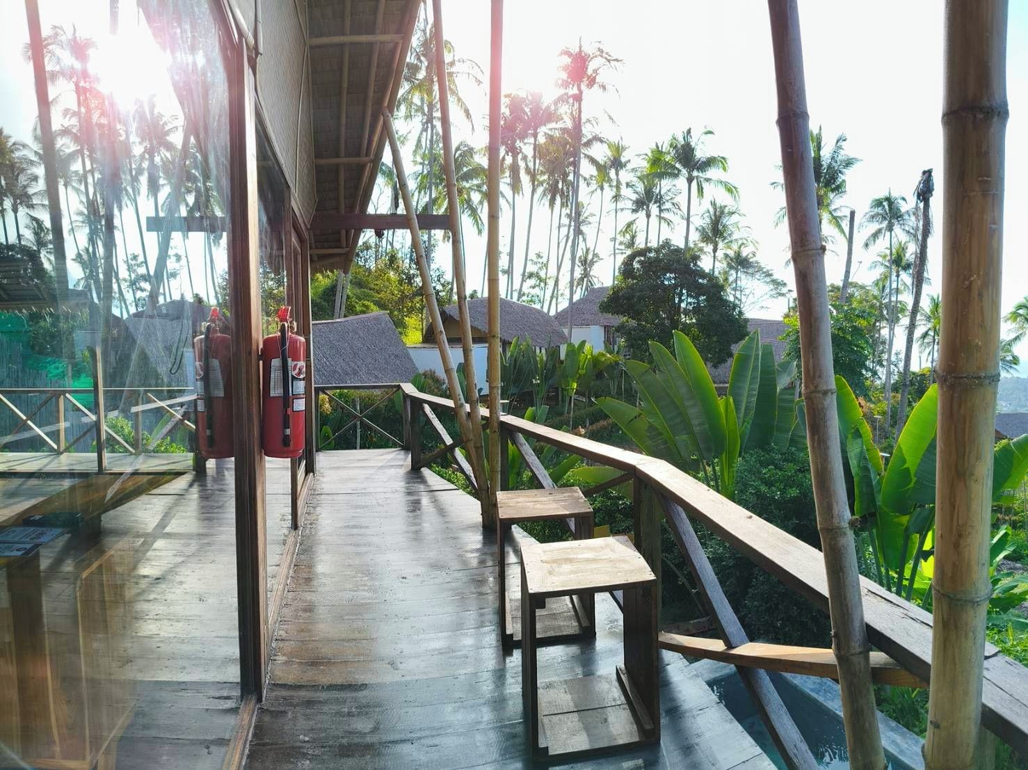 A wooden deck with a fire extinguisher, a bench, and a view of palm trees and thatched-roof buildings.