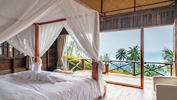 A bedroom with a four-poster bed, a balcony with a view of palm trees and the ocean, and wooden flooring. - RoomQuest THEVA Resort koh Samui  (Koh Samui)