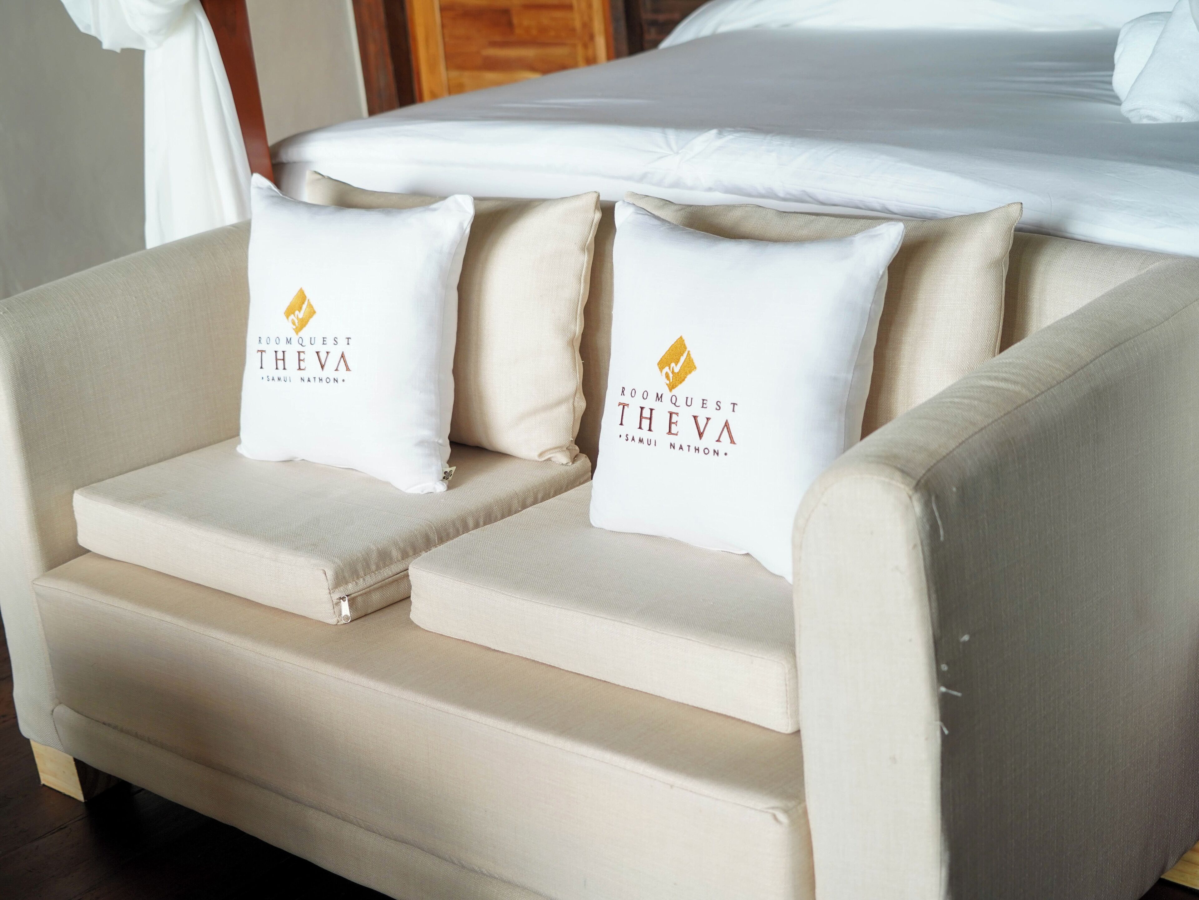 A beige sofa with two pillows featuring the RoomQuest Theva logo.