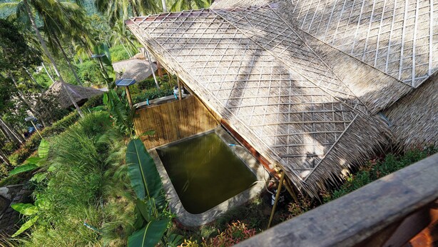 Estructura con techo de paja, fuente de agua y abundante vegetación.