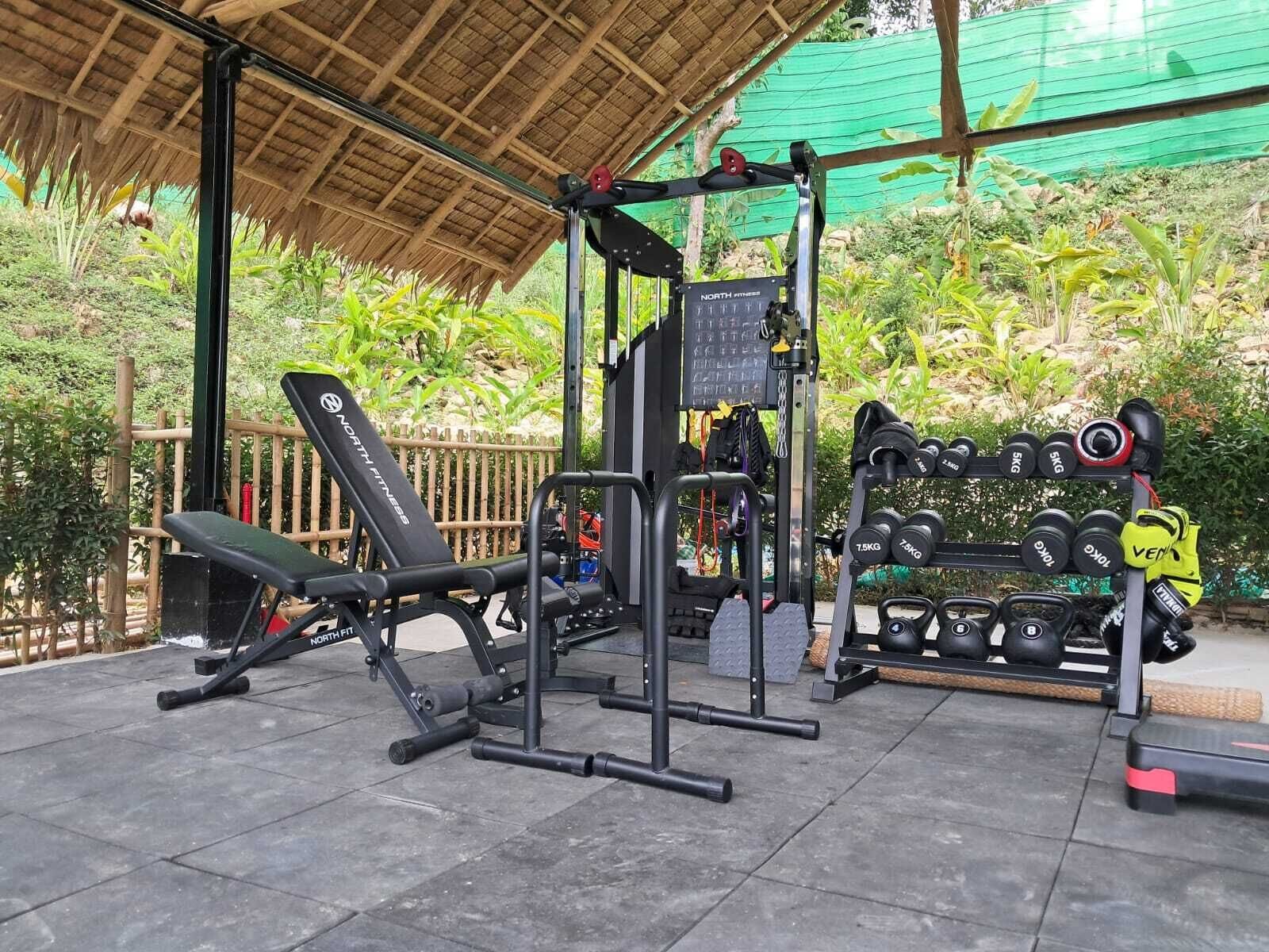 A covered outdoor gym area with exercise equipment, including a bench, squat rack, and dumbbells.