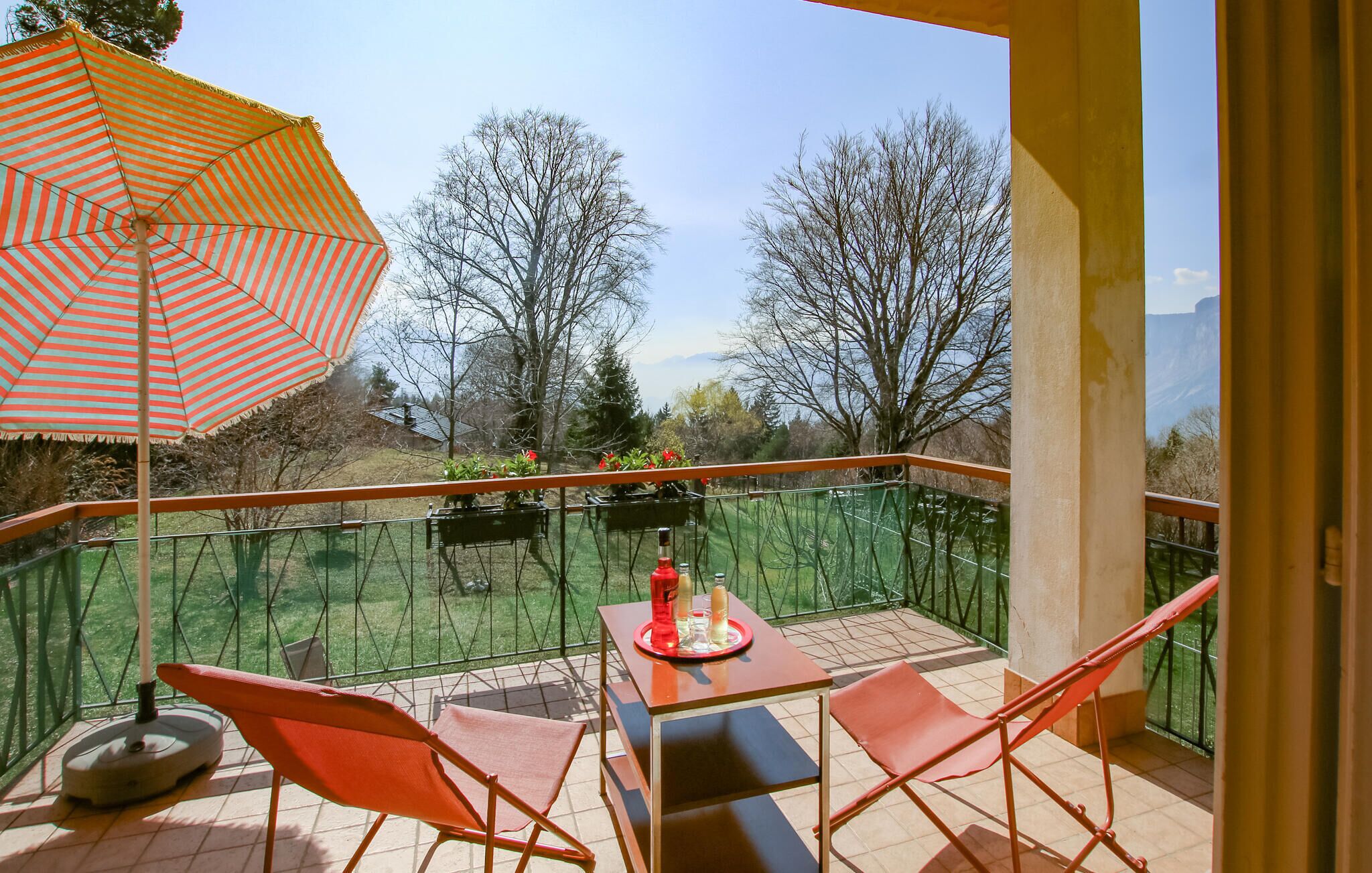 A balcony with two red chairs, a table with a tray of items, and a striped umbrella.