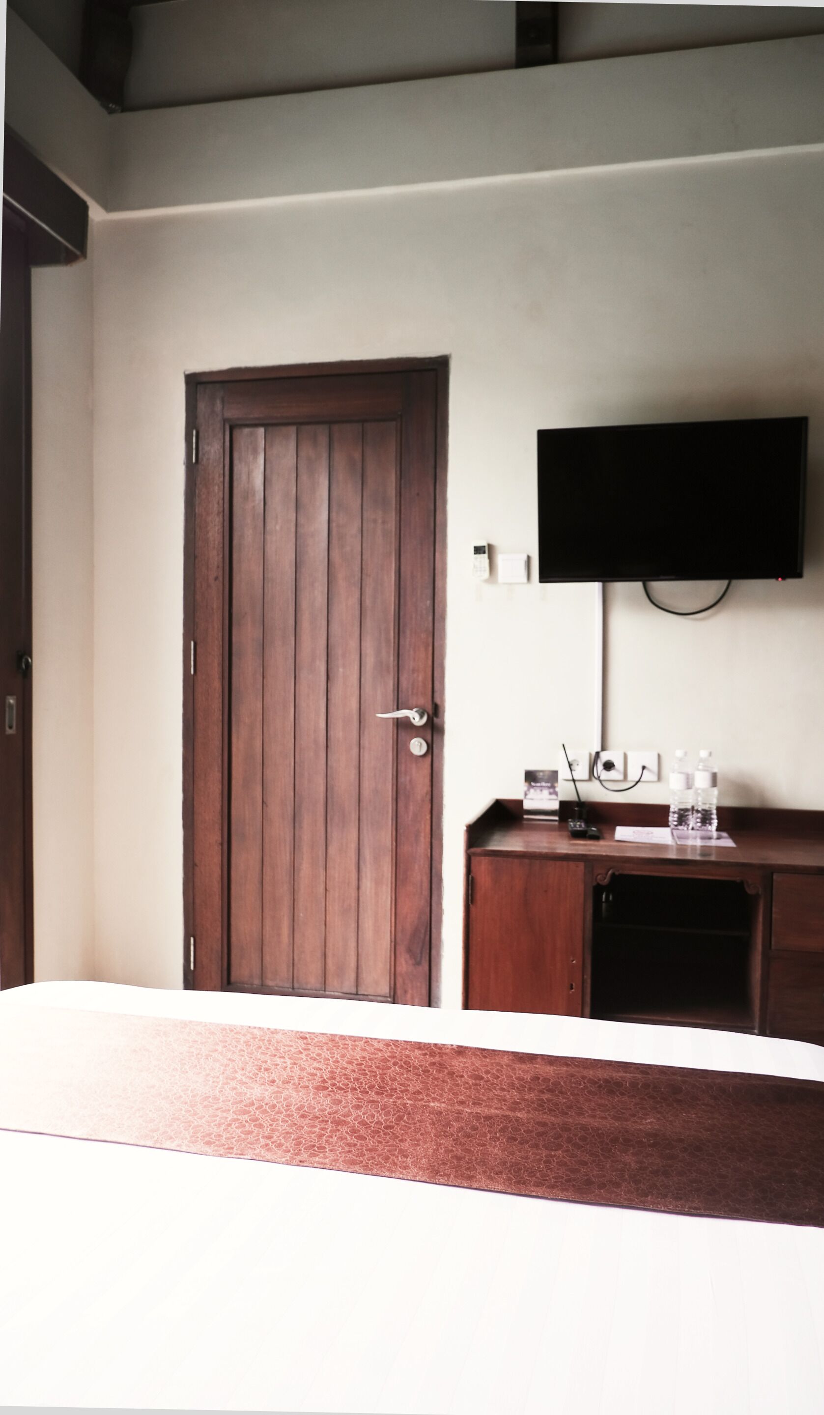 Une pièce avec une porte en bois, un téléviseur à écran plat fixé au mur, une petite armoire en bois et un lit avec une tête de lit blanche.