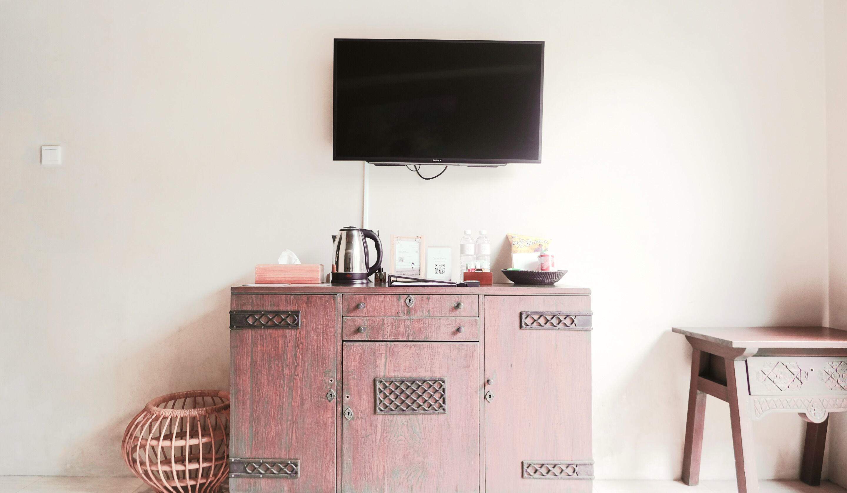 A room with a wooden cabinet, a television mounted on the wall, a wicker chair, and a small table.
