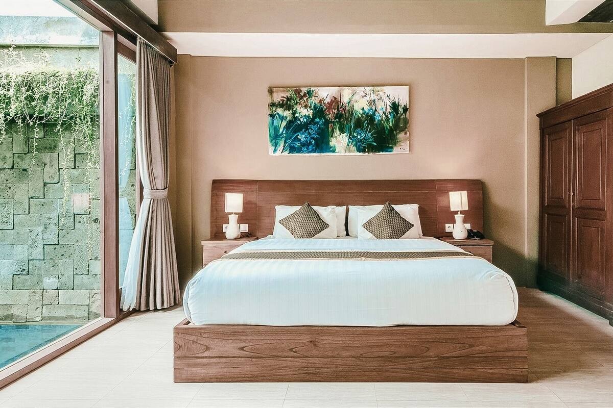 A bedroom with a large bed, a painting on the wall, and a view of a pool.