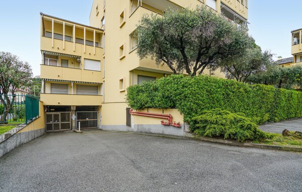 Un edificio amarillo de varios pisos con entrada protegida por una reja, un árbol y un seto.