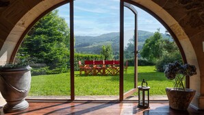 Une pièce dotée d’une grande fenêtre en arc d’où l’on peut admirer un paysage verdoyant, avec une table et des chaises rouges disposées à l’extérieur.