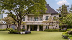 Un edifici gran de dos pisos amb una entrada formal i un jardí ben cuidat amb gespa, arbres i una banca.