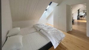 A bedroom with a sloped ceiling, a skylight, a bed with white bedding, and wooden flooring.