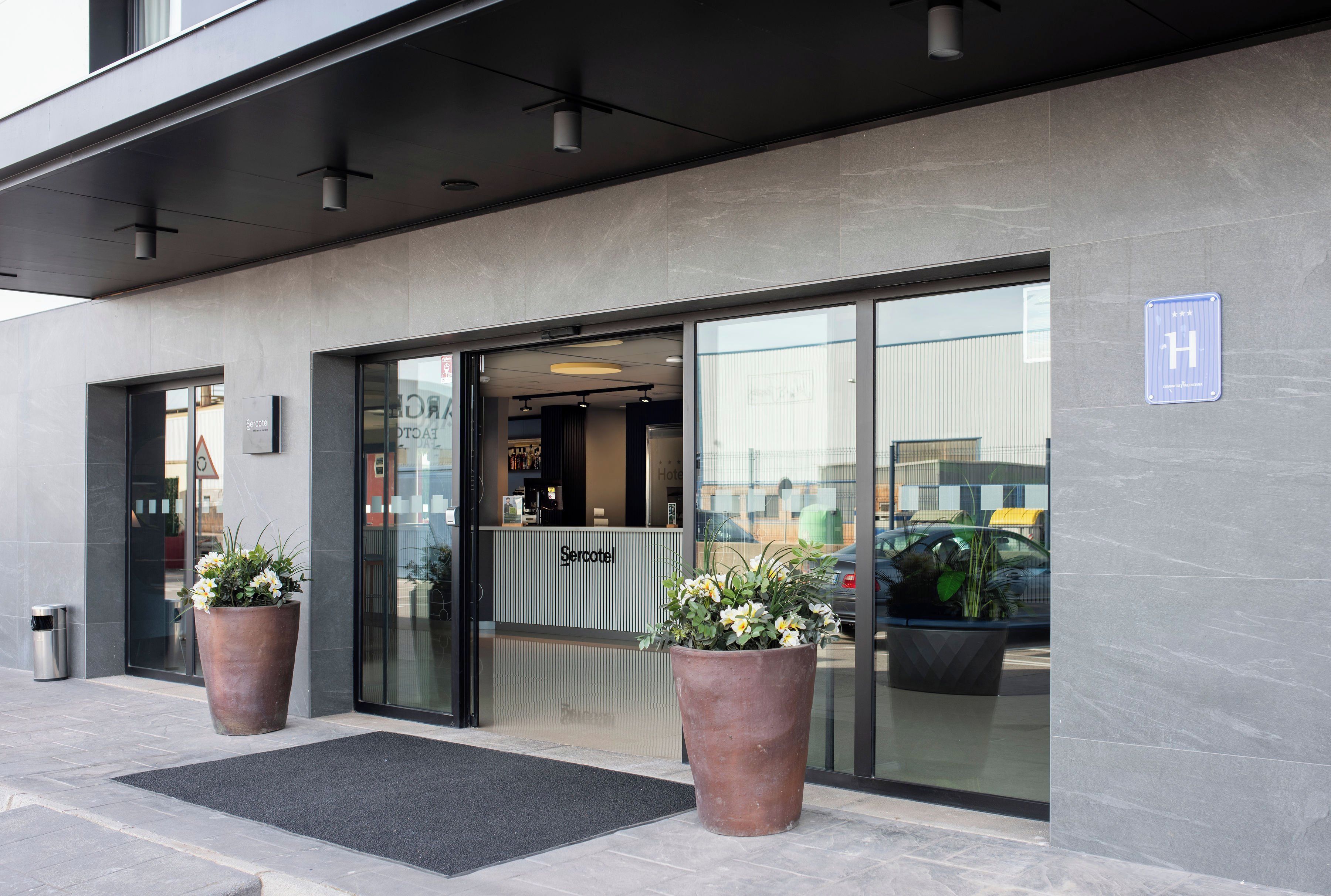 Entrada de un hotel con un mostrador de recepción, plantas en macetas grandes y una vista despejada del interior a través de puertas de vidrio.
