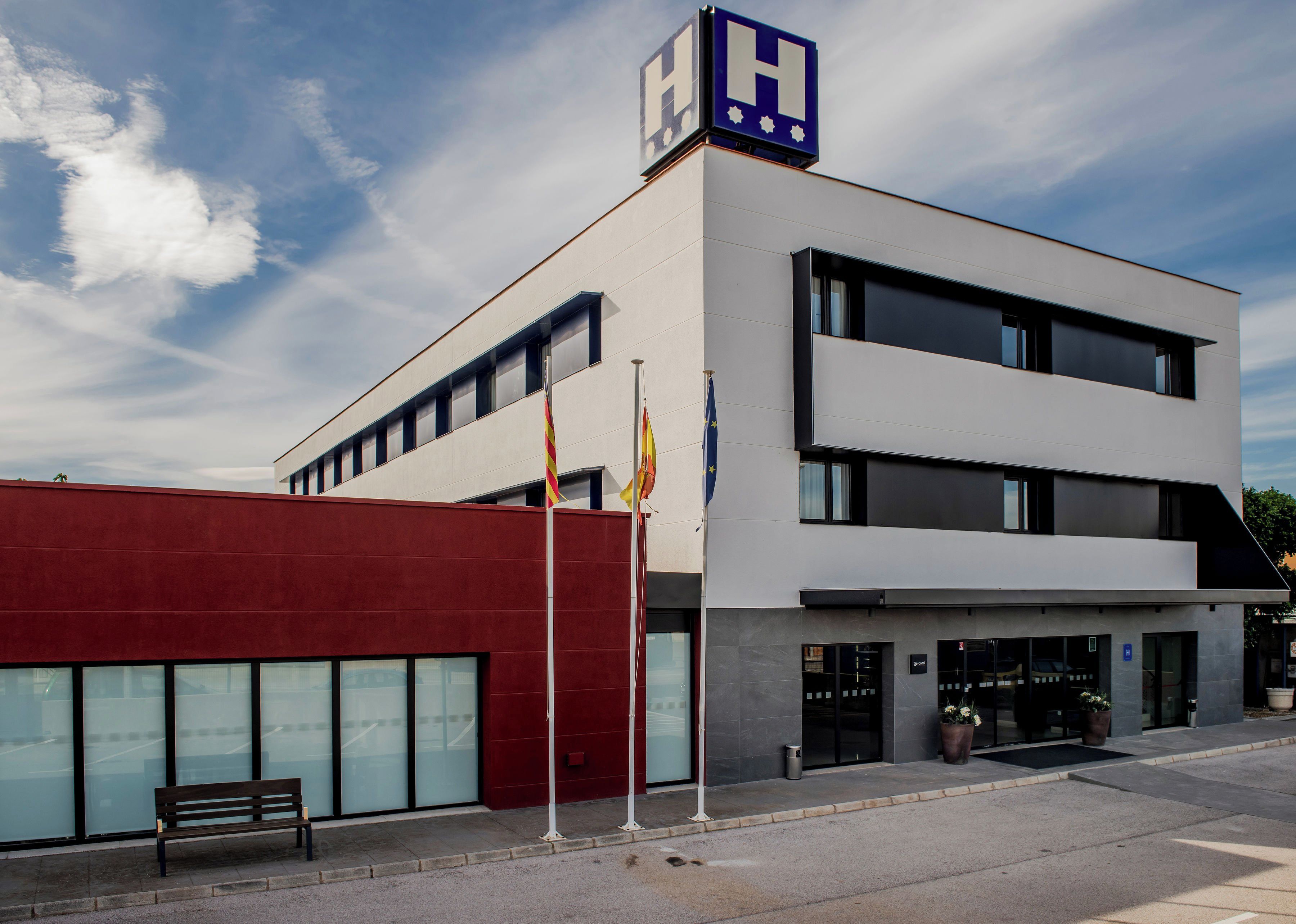 Un edificio moderno con un llamativo letrero con la letra 'H', indicando un hotel. Hay banderas en los mástiles, un banco y un área de entrada pavimentada.