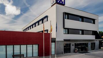 Un edificio moderno con un llamativo letrero con la letra 'H', indicando un hotel. Hay banderas en los mástiles, un banco y un área de entrada pavimentada.