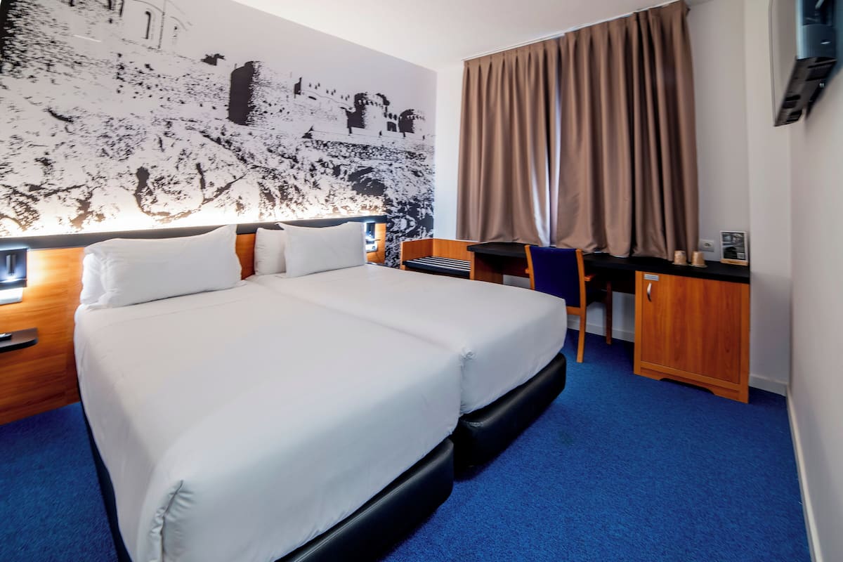 Quarto de hotel com uma cama grande, uma escrivaninha, uma cadeira e uma televisão presa à parede.