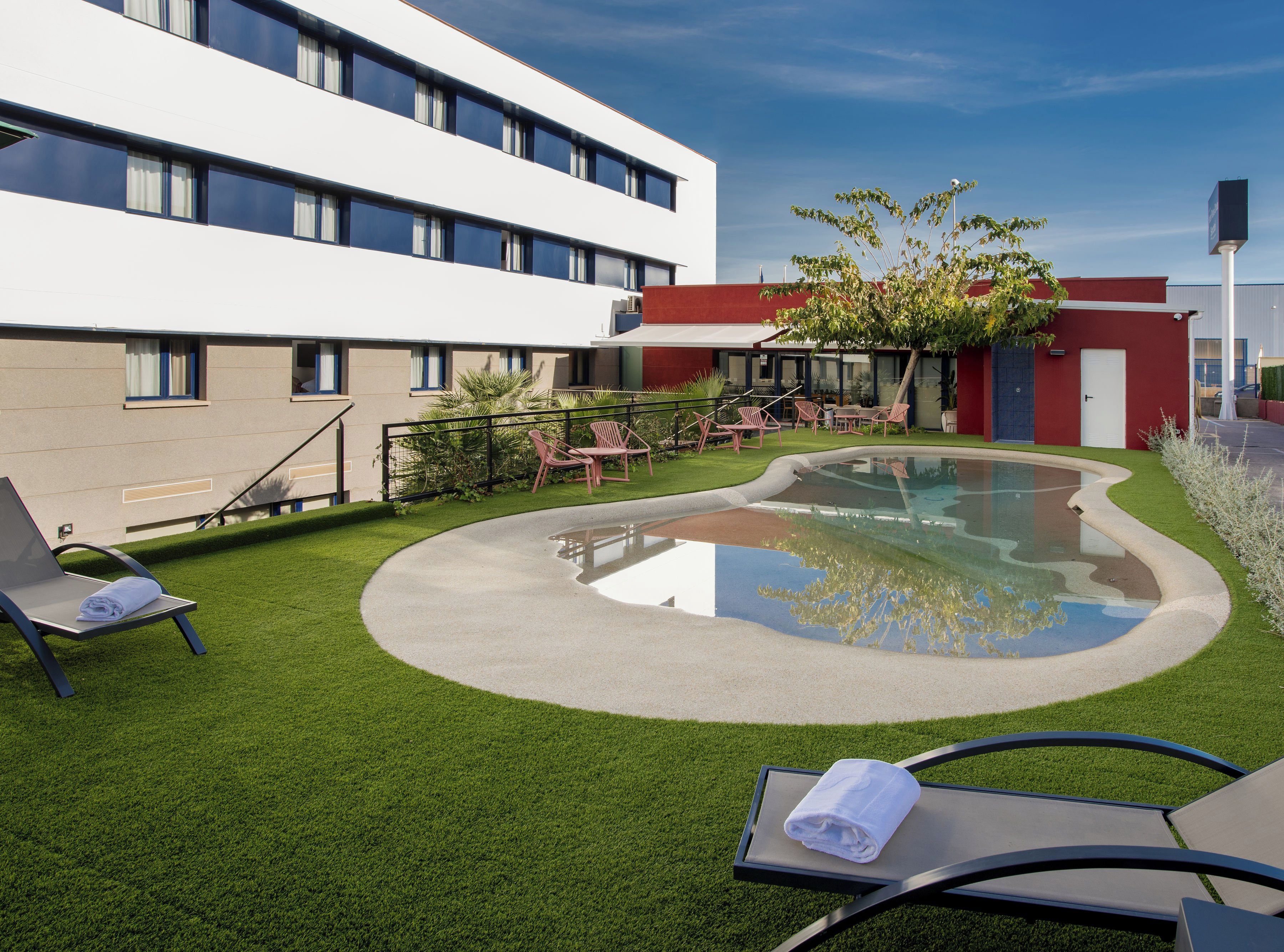A modern hotel courtyard with a swimming pool, lounge chairs, and a well-maintained lawn.