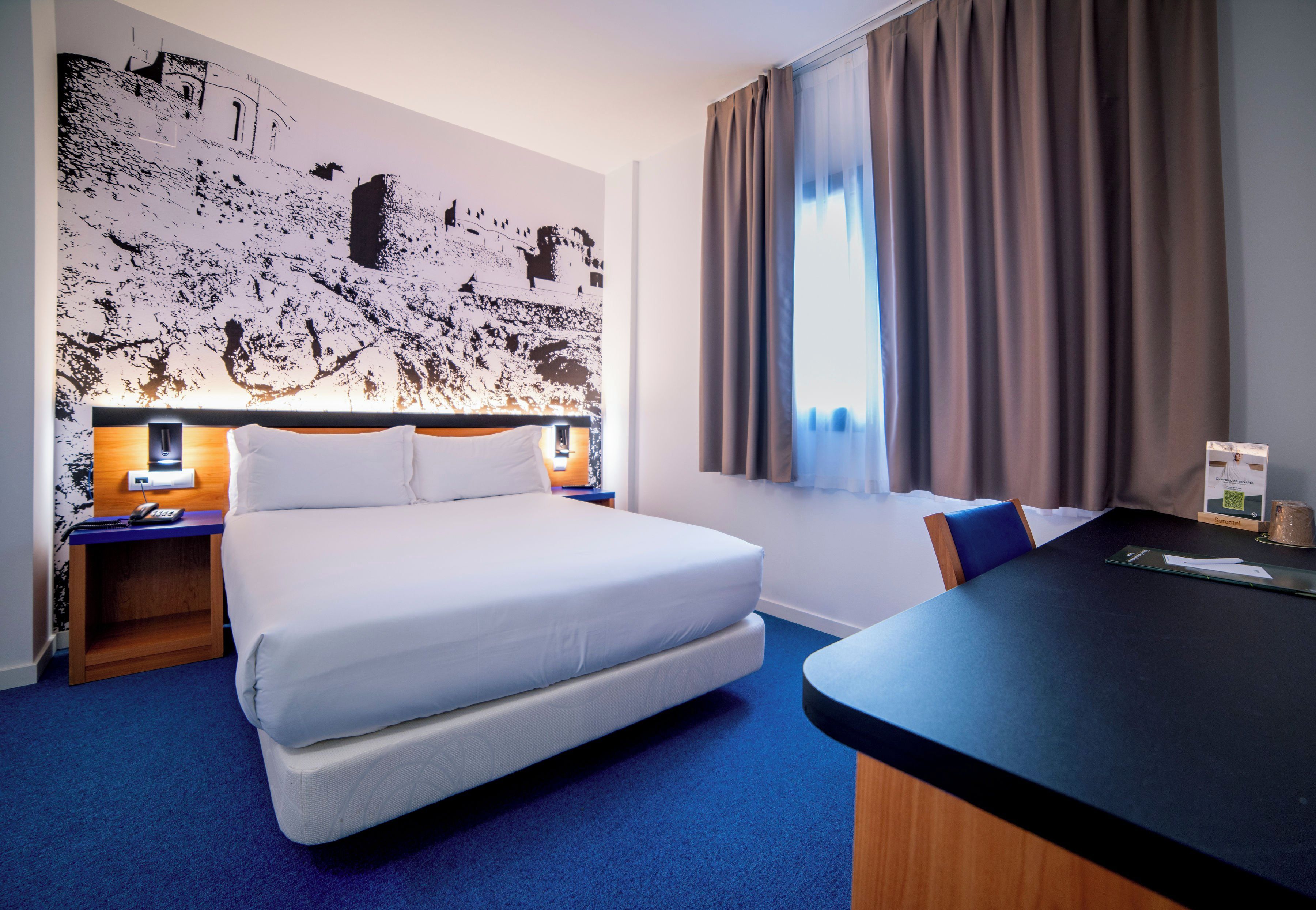 A hotel room with a large bed, a desk, a chair, a nightstand, a telephone, and a wall with a textured design.