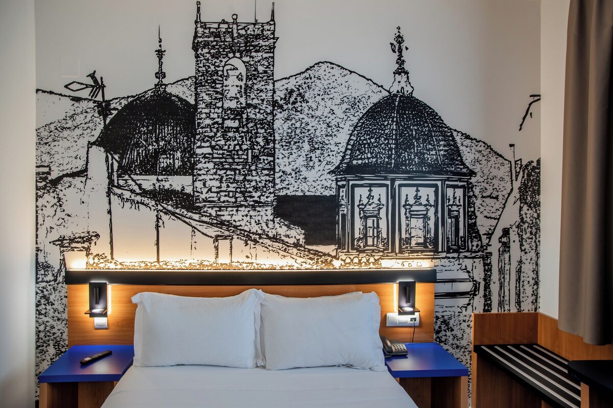 A hotel room with a bed, a nightstand, a wall mural of a cityscape, and a wooden headboard.