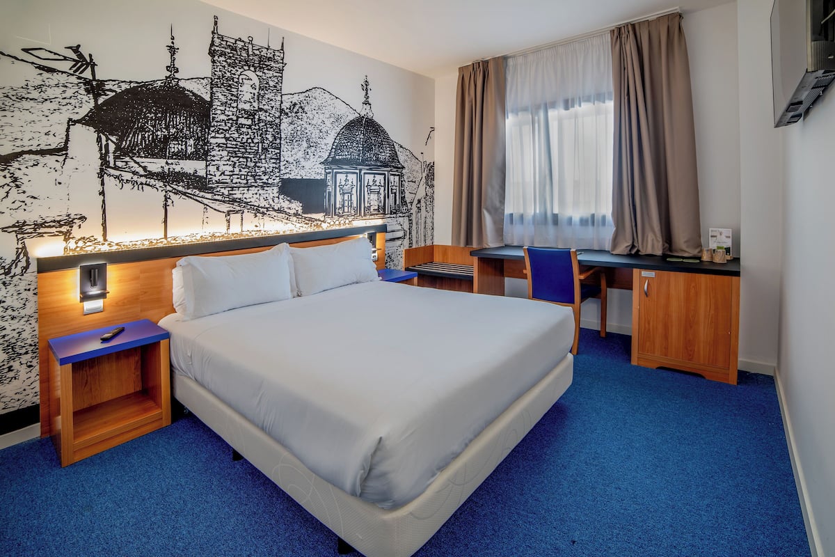 Quarto de hotel com uma cama, uma escrivaninha e um mural na parede.