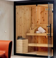 Una sauna con paredes de madera y puerta de vidrio, un banco de madera, una toalla y un cubo con piedras.