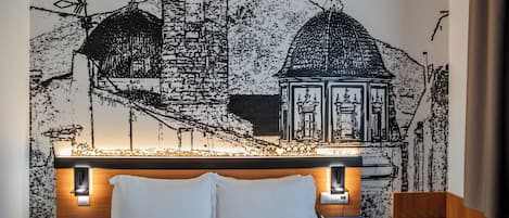A hotel room with a bed, bedside tables, a wall-mounted lamp, and a decorative wall mural.