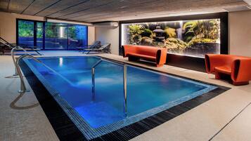 Un área de piscina interior moderna con agua cristalina, sillas de descanso y un amplio ventanal con vistas a un paisaje rocoso.
