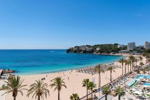 A beach with a pool area, palm trees, and buildings in the background.