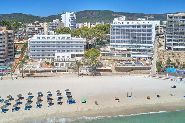 Un hotel frente a la playa con varios pisos, un área de piscina y sombrillas.