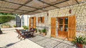 Un edificio de piedra con contraventanas de madera, un porche cubierto con una mesa y sillas, y plantas en macetas.