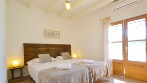 Un dormitorio con dos camas, un cabecero de madera, una lámpara de noche, un cuadro enmarcado en la pared y un amplio ventanal con cortinas.