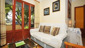 Ein Wohnzimmer mit einer Couch, einem Glastisch und einer Tür, die auf einen Balkon führt. An den Wänden befinden sich Bilder in Rahmen und auf einem Beistelltisch steht eine Lampe.