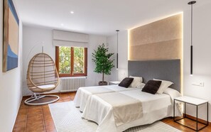 A modern bedroom with two beds, a hanging chair, a potted plant, and a view of greenery outside.