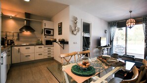 Cocina moderna con armarios blancos, microondas y estufa. Hay un comedor con mesa de vidrio y sillas, y vistas al exterior a través de un amplio ventanal.