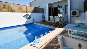 Un área de piscina moderna al aire libre, con una piscina azul cristalina, una terraza de madera y una mesa con sillas.