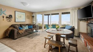 A living room with a sofa, coffee table, and chairs, a fireplace, and a large window with a view of the beach. - Ultimate Oceanfront Escape for Two at Beachy Eagle B, Amazing Views, Fire-Table, Steps from Beach (Rockaway Beach)