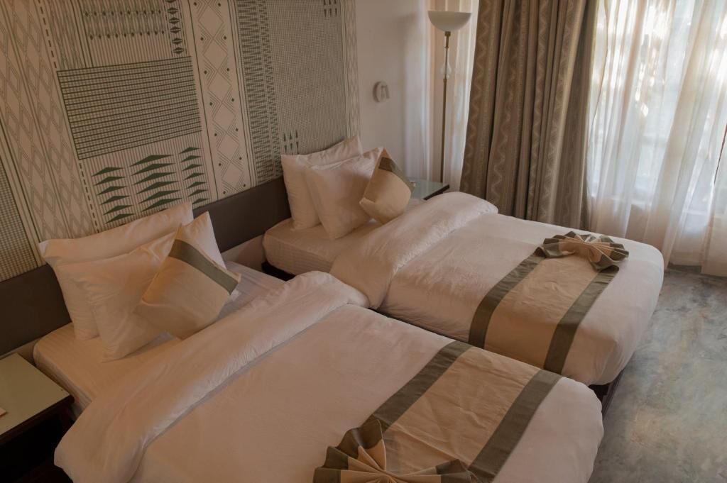 A hotel room with two beds, a patterned wall, a lamp, and a window with curtains.