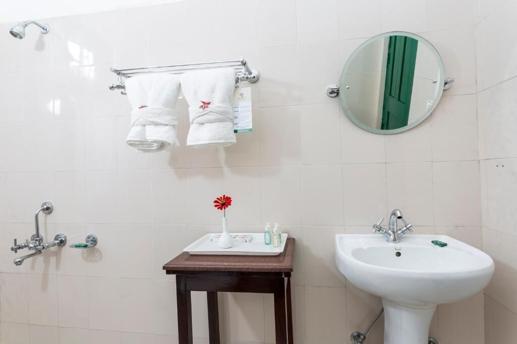 Casa de banho com lavatório branco, espelho, porta-toalhas com duas toalhas, pequena mesa com artigos de higiene e chuveiro.