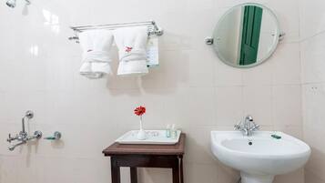 Casa de banho com lavatório branco, espelho, porta-toalhas com duas toalhas, pequena mesa com artigos de higiene e chuveiro.