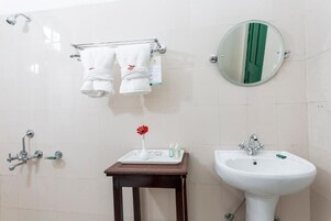 Casa de banho com lavatório branco, espelho, porta-toalhas com duas toalhas, pequena mesa com artigos de higiene e chuveiro.