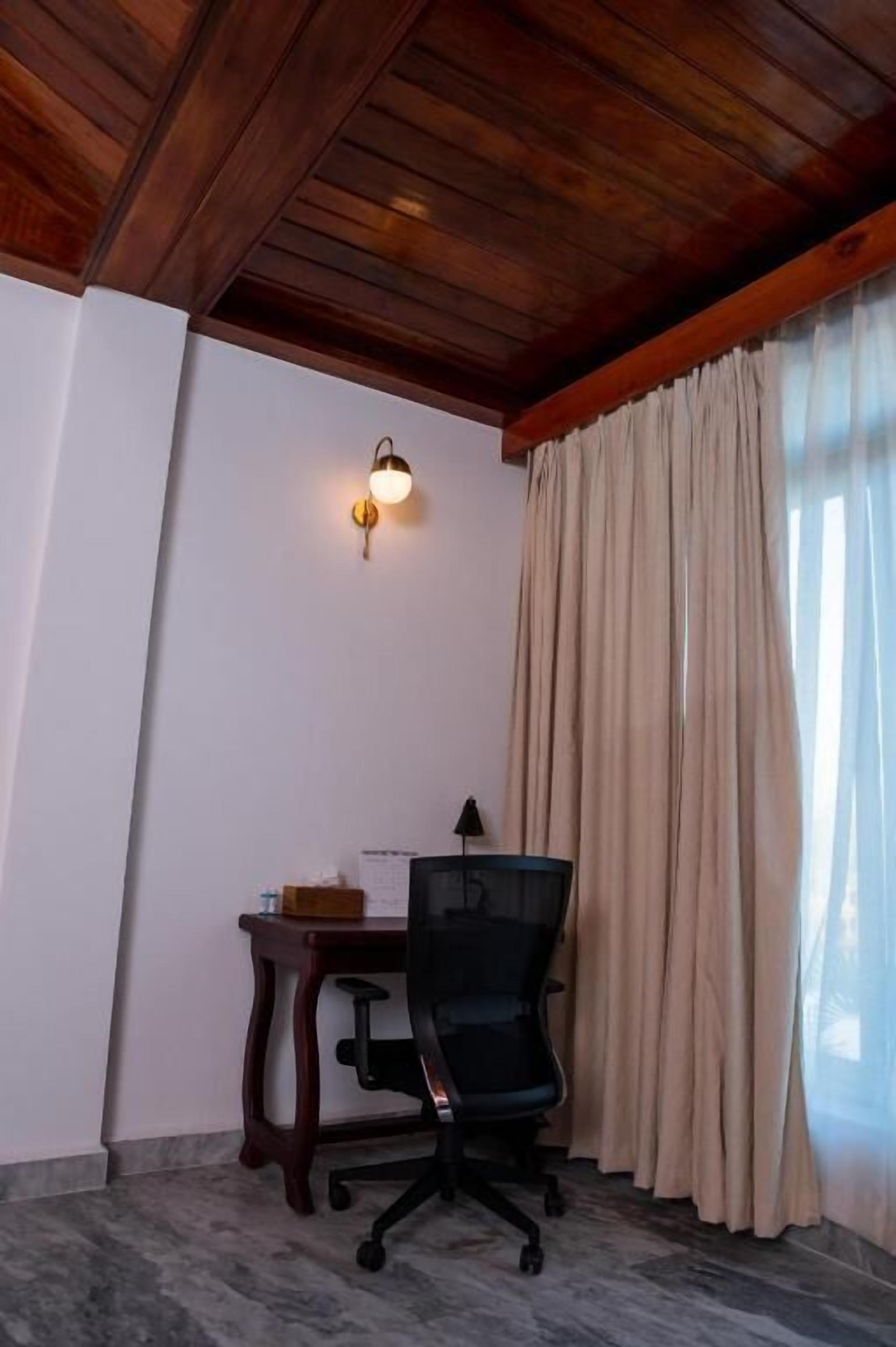 A room with a wooden ceiling, a desk with a chair, a wall-mounted light, and a window with curtains.