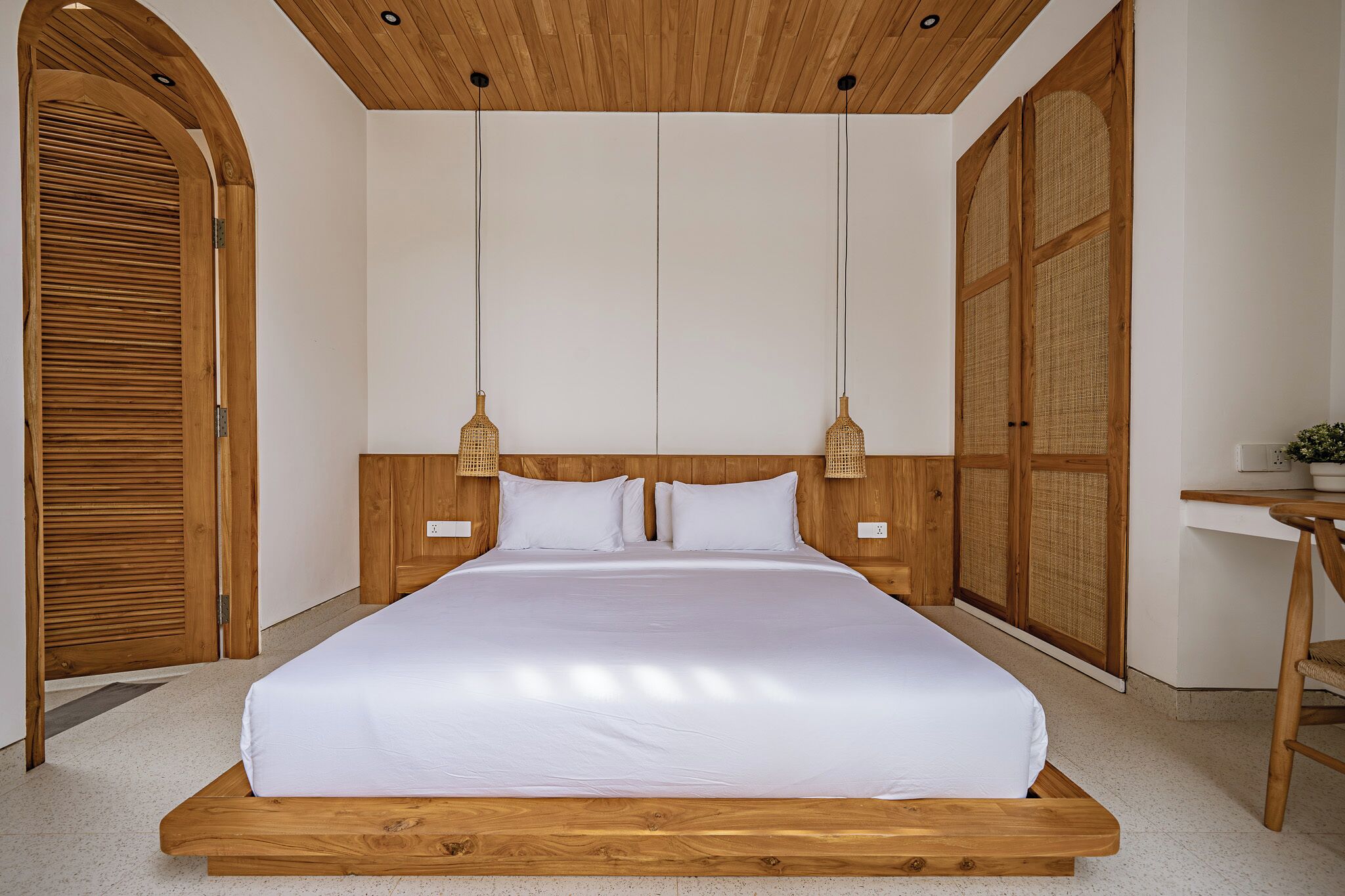 Ein großes Bett mit Holzrahmen und weißer Bettwäsche, zwei hängende Leuchten und ein Schreibtisch mit Stuhl.