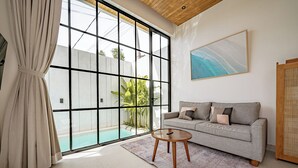 Een moderne woonkamer met een grote schuifpui naar een zwembad, een grijze bank met kussens, een klein houten bijzettafeltje en een ingelijste afbeelding aan de muur.